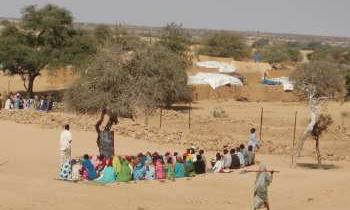 A group of children during a lesson outdoors, sat on the ground beside a tree.